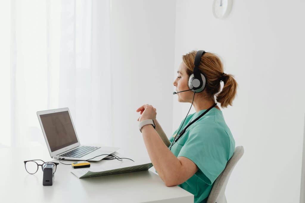 A person in a teal medical uniform wearing headphones, sitting at a desk with a laptop, glasses, and a small device. Soft natural light filters in.
