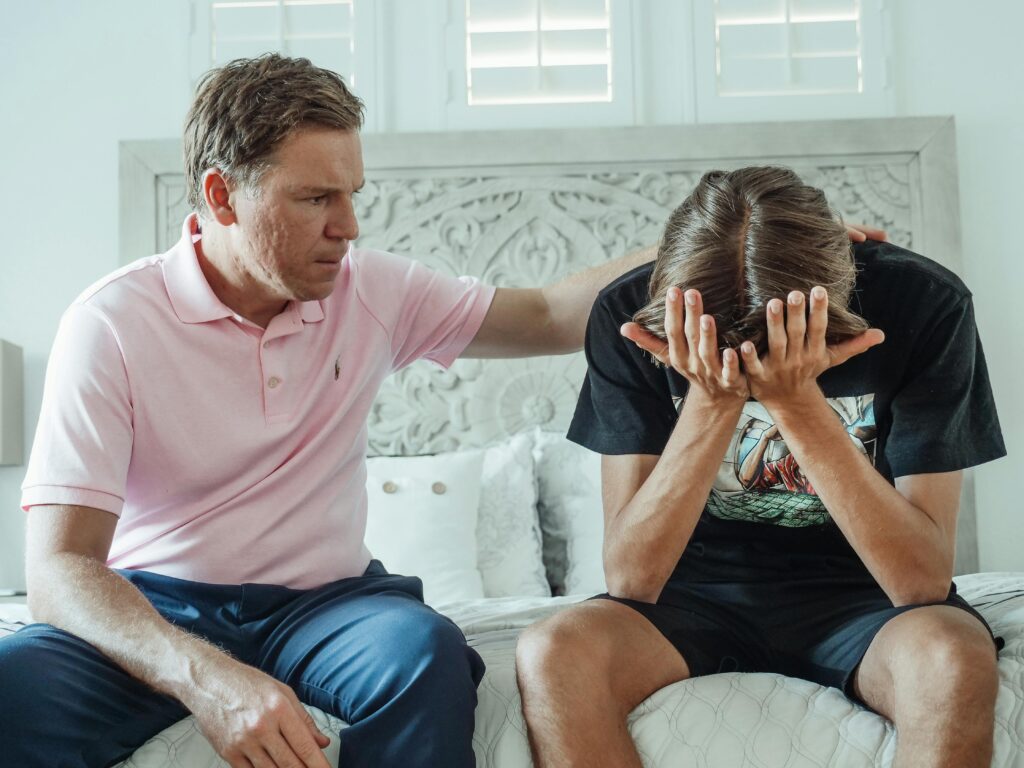 A concerned adult sits beside a young person, who is holding their head in distress, suggesting a moment of emotional support in a comforting setting.