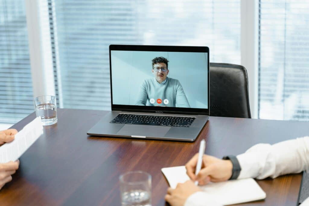 A video call in a modern office setting, with a laptop displaying a person on screen, and two individuals taking notes.