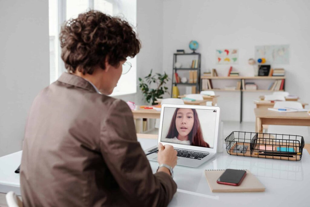 A person in a blazer is engaged in a video call on a laptop at a bright classroom, with desks and shelves in the background.
