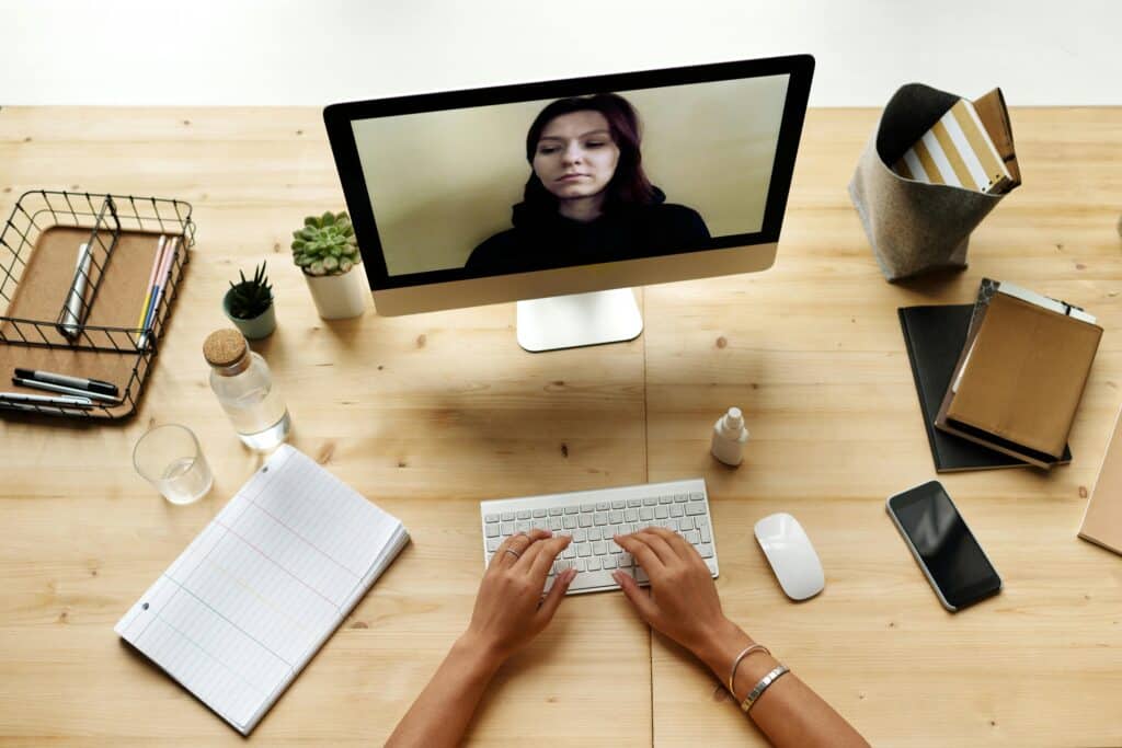 Overhead view of a person at a desk during a video call. The desktop has a keyboard, a notepad, a phone, and small plants, conveying a work-from-home vibe.