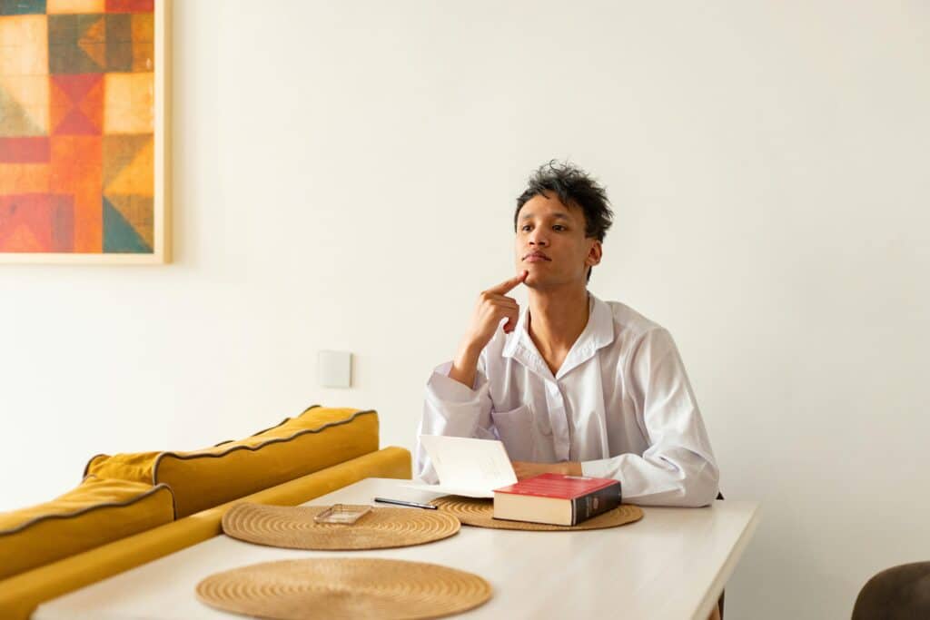 A person sits at a table with a notebook and a stack of books, deep in thought, surrounded by a bright, minimalist room.