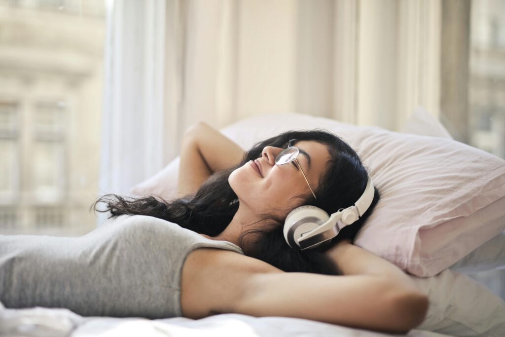 A person relaxes on a bed, wearing white headphones, with arms behind their head, surrounded by soft pillows and natural light.