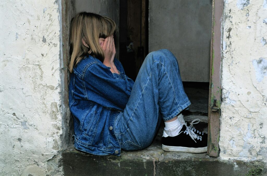 A girl in a denim jacket and jeans sits in a doorway, face covered with her hands, conveying sadness or distress in a stark setting.