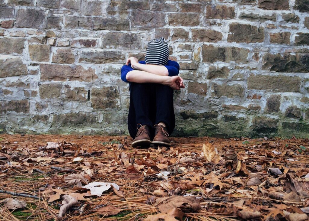 A person sits against a stone wall, hugging their knees, surrounded by autumn leaves, wearing a striped beanie.