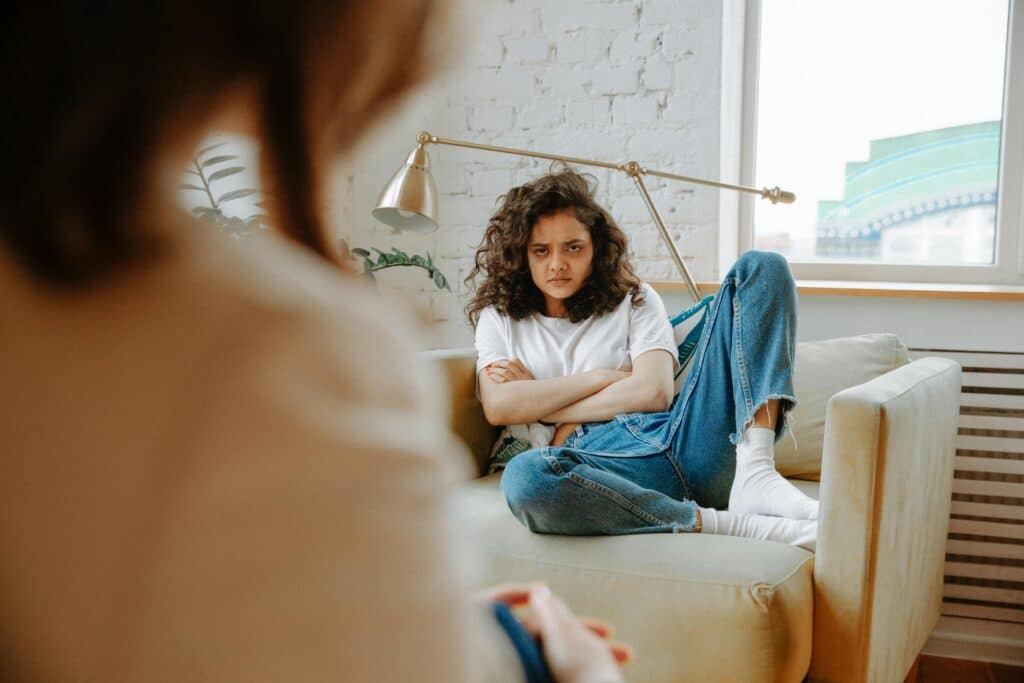 A person with curly hair sits with arms crossed on a couch, facing a large window in a cozy, well-lit room.