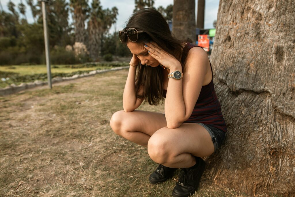 A woman squats beside a large tree wearing a striped tank top, denim shorts, and black boots, appearing deep in thought.