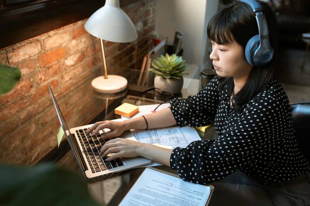 A person types on a laptop at a wooden desk, surrounded by notes, a plant, and a lamp, in a cozy workspace with exposed brick walls.