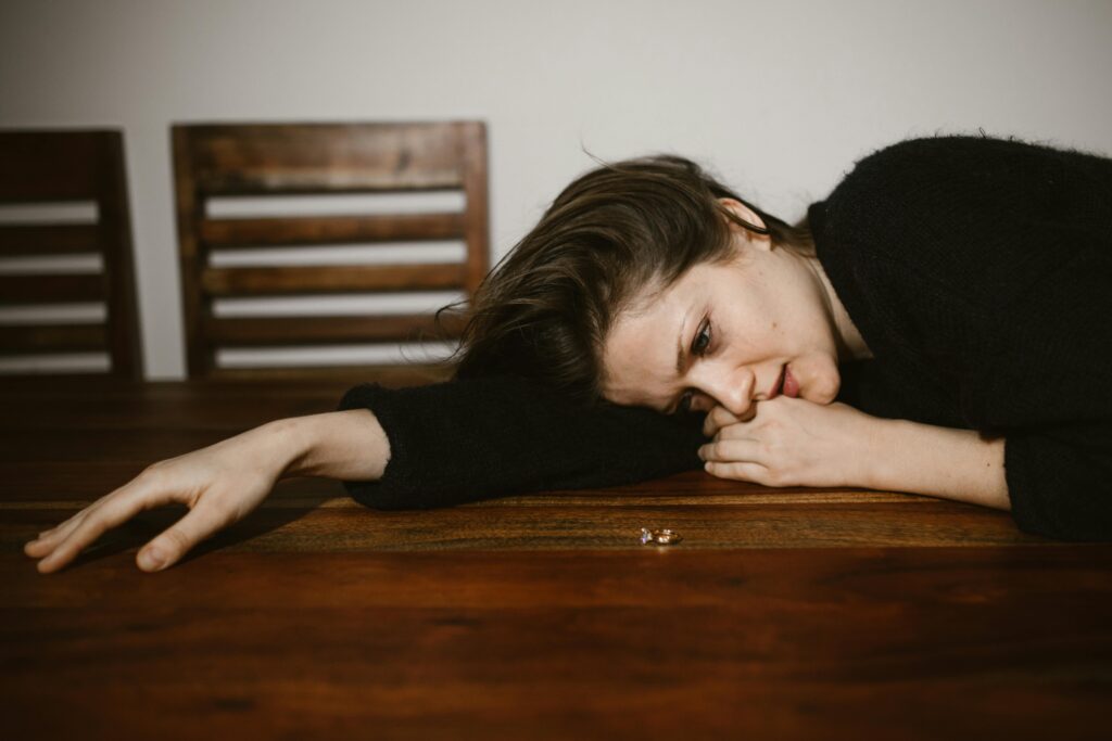 A person lies on a wooden table, with an outstretched arm and two rings on the surface, evoking a sense of despair or loss.