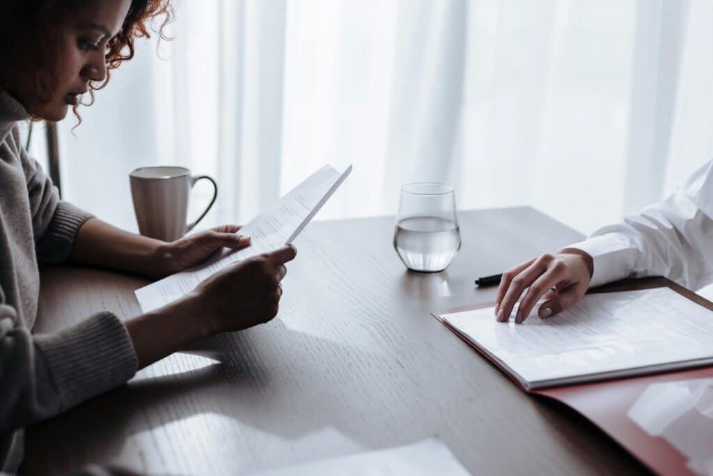 A hand holding documents sits at a table with a cup and a glass of water, illuminated by soft sunlight through sheer curtains.