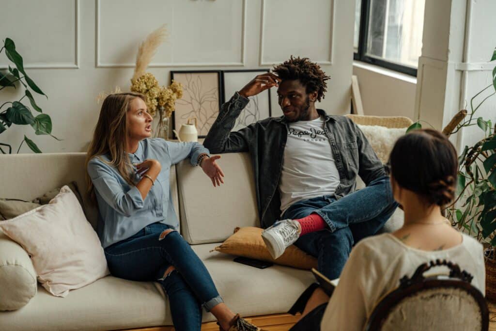 A group of three people seated on a couch in a cozy, well-lit room, engaging in conversation with plants and artwork in view.