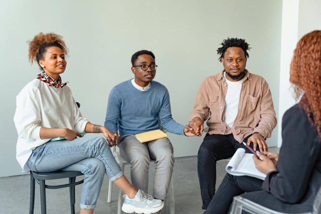 A group of four people sits in a circle, holding hands, engaged in a supportive discussion, with a neutral backdrop.