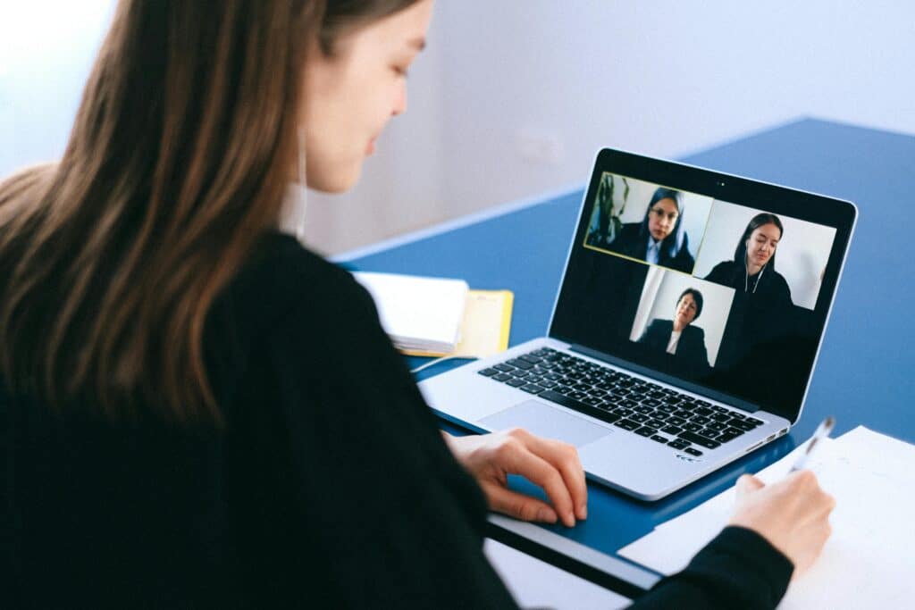 A woman in a black sweater participates in a video conference on a laptop with three colleagues visible on the screen, set on a blue table.