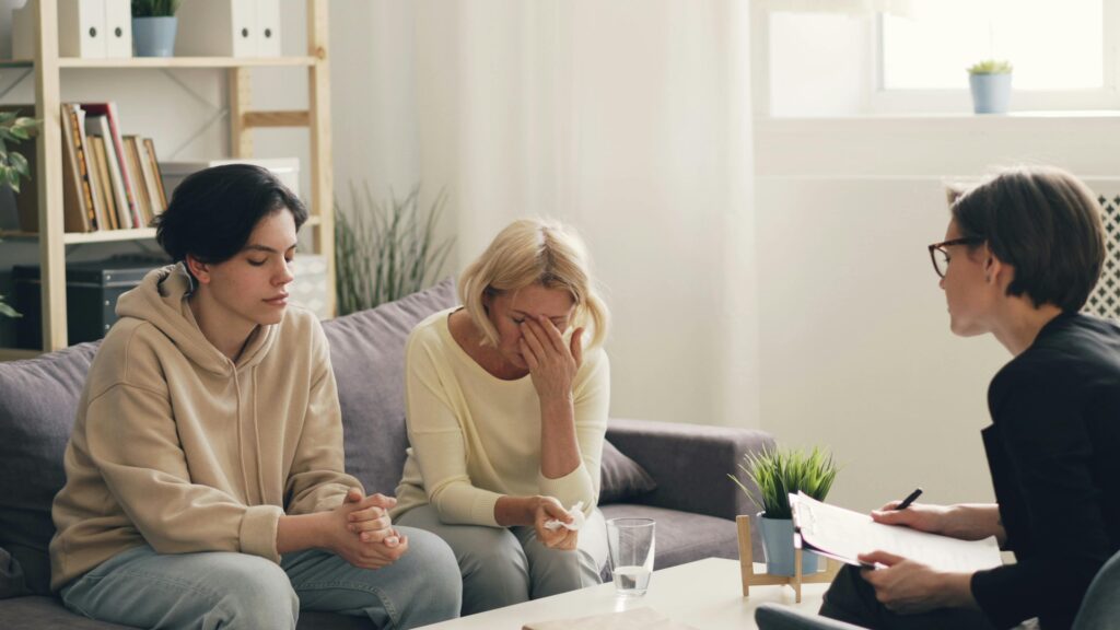 Two individuals sit on a couch in a bright room, listening attentively to a woman with a clipboard during a discussion or therapy session.