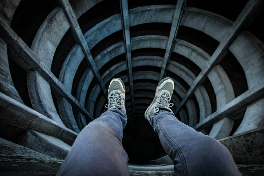 A person's feet, wearing sneakers, dangle over the edge of a spiral concrete structure, emphasizing a sense of height and depth below.