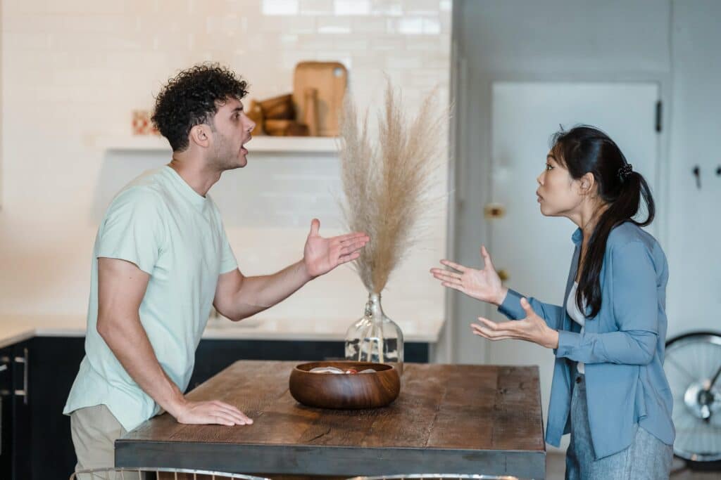 A man and woman are engaged in an animated discussion at a wooden table, with a vase of dried grass in the background.