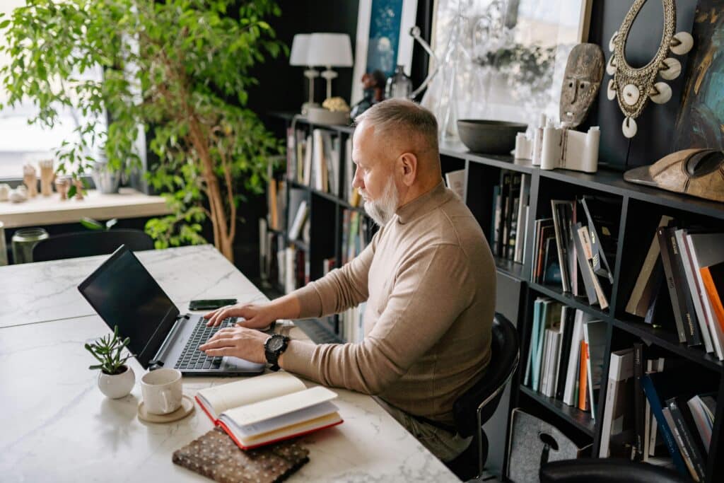 A person types on a laptop at a stylish desk surrounded by plants and bookshelves, with notebooks and cups nearby.