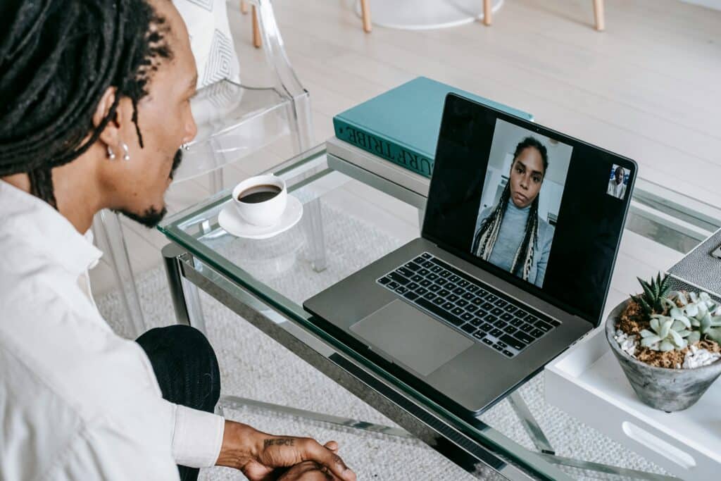A person with dreadlocks sits at a glass table, engaged in a video call on a laptop, with a cup of coffee nearby.