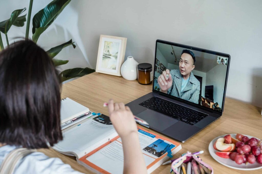 A person engages in a video call on a laptop, surrounded by study materials, snacks, and a plant on a wooden desk.