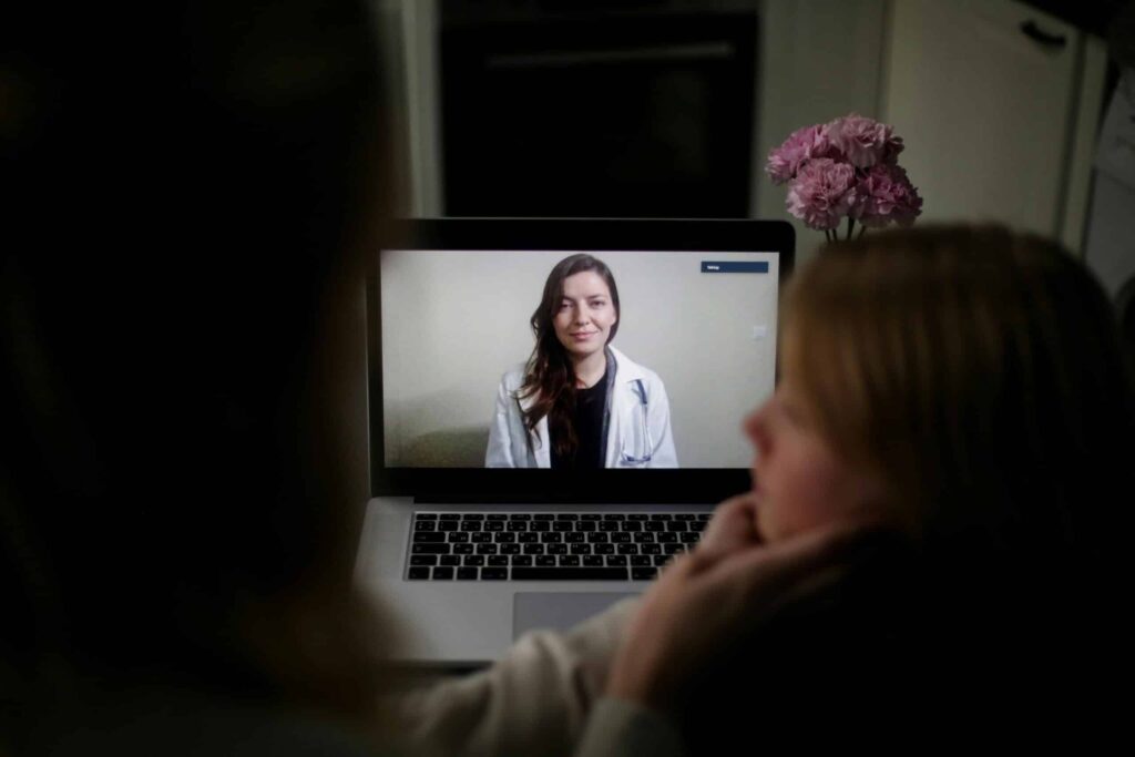 A laptop displays a healthcare professional speaking via video call, while a person watches from the side, with flowers in the background.