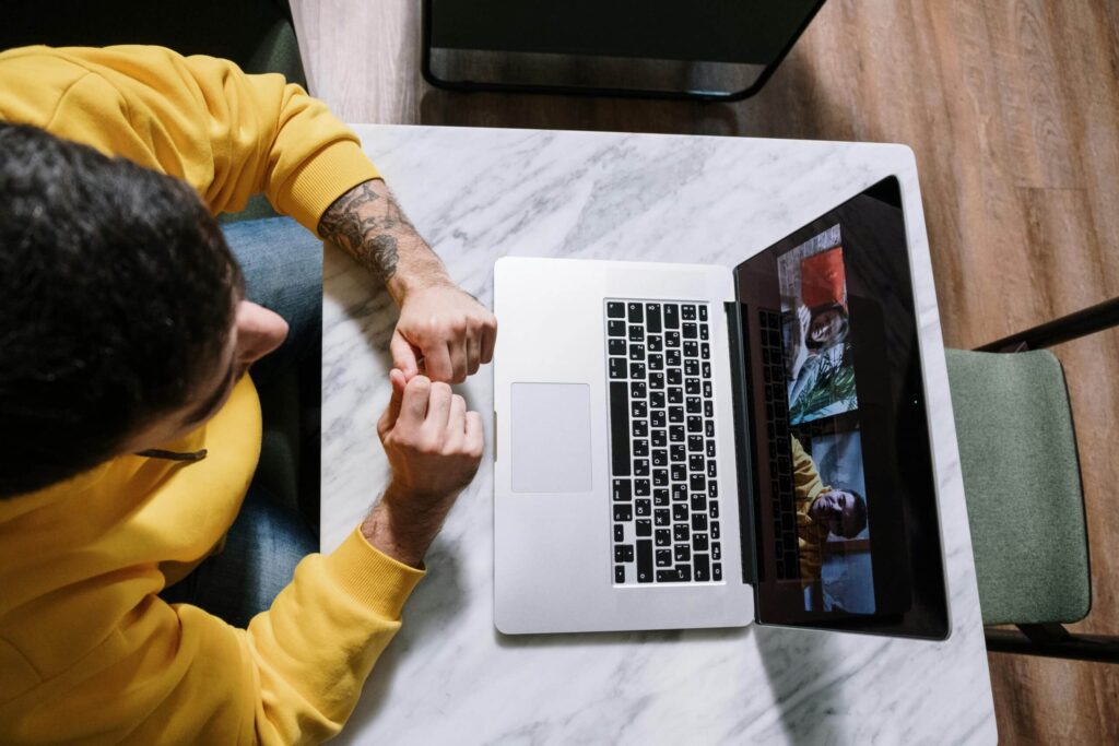 A man in a yellow hoodie watches a video call on his laptop, resting his hands on a marble table.