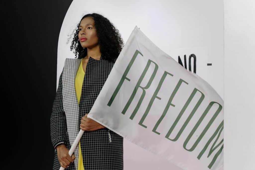 A person in a checkered jacket holds a large white flag with the word "FREEDOM" in green, against a minimalist backdrop.
