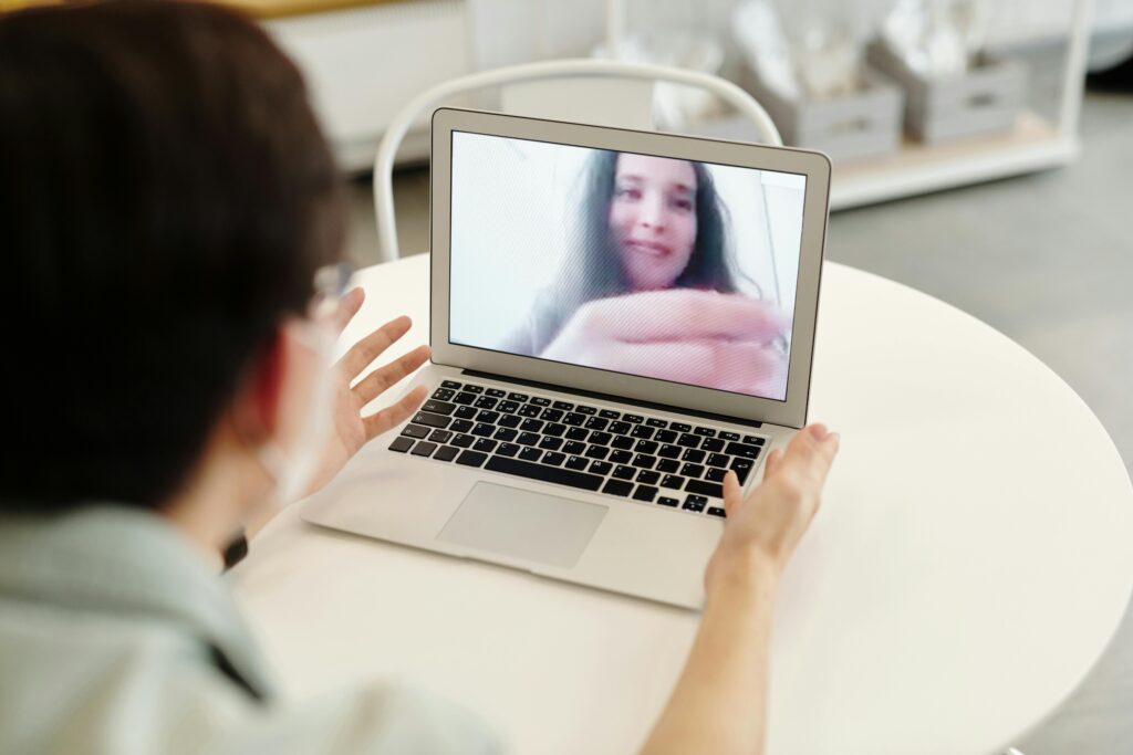 A person sits at a table, engaging in a video call on a laptop, gesturing with hands, indicating interaction or discussion.