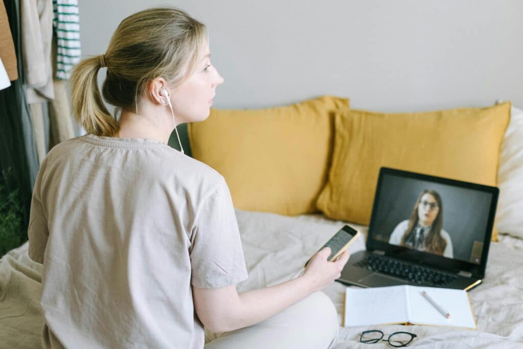 A person sits on a bed with a laptop and phone, engaged in a video call, surrounded by cozy yellow pillows and a notepad.