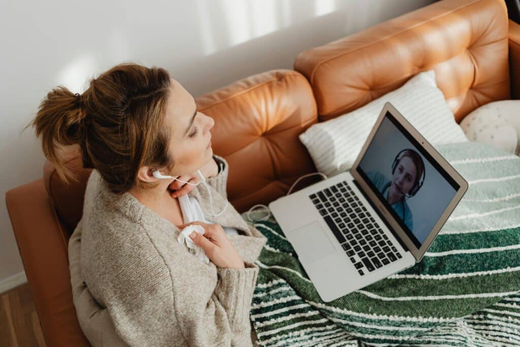A woman with earbuds, wrapped in a green-striped blanket on a brown couch, engaging in a video call on a laptop. The scene is warm and relaxed.