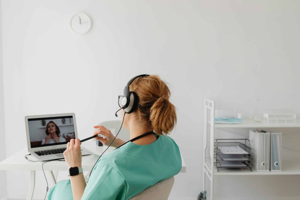 A healthcare professional in scrubs uses a headset while consulting on a laptop in a bright, minimalistic office space.