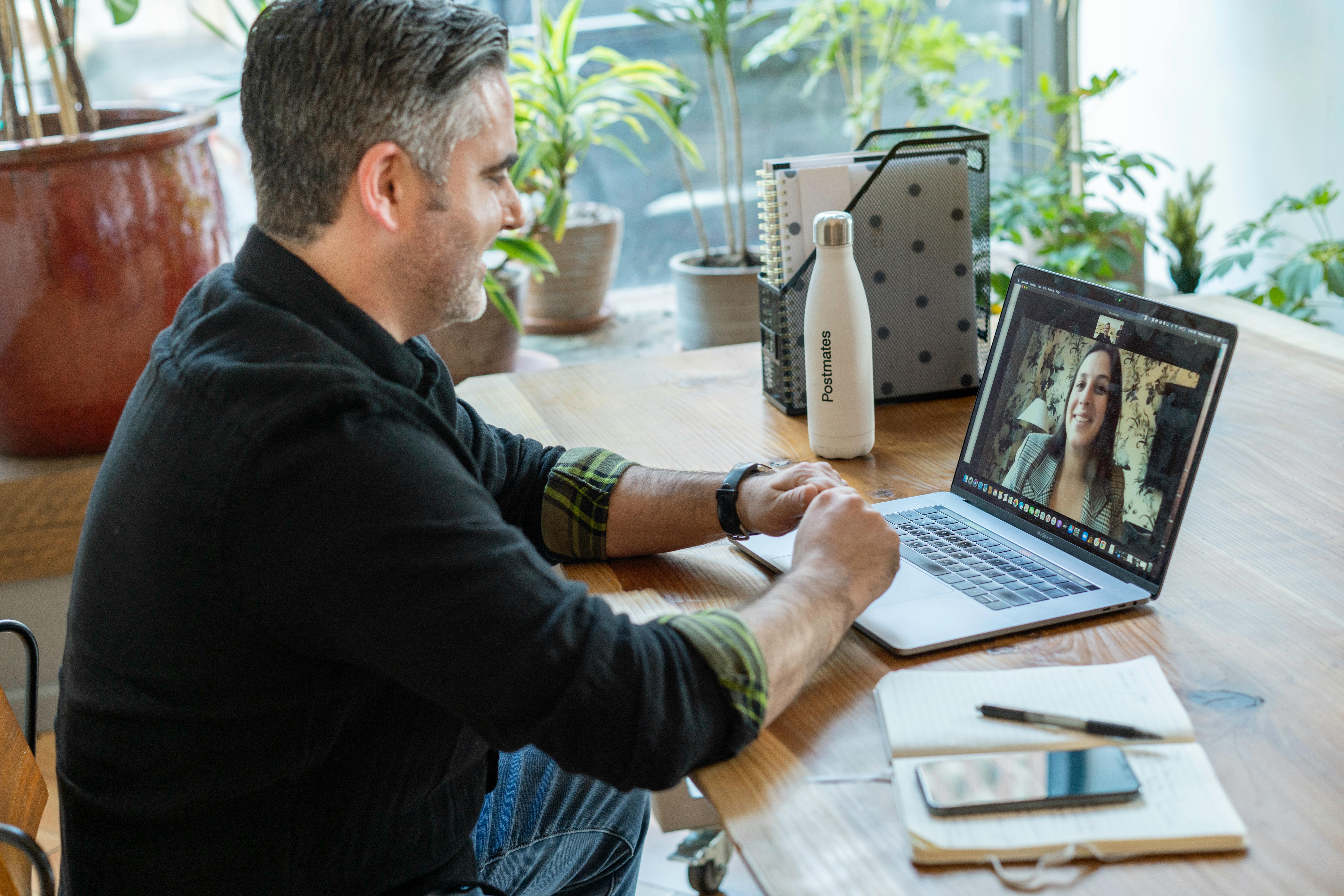 A man works at a wooden desk with a laptop, notebook, and water bottle, engaging in a video call surrounded by plants.