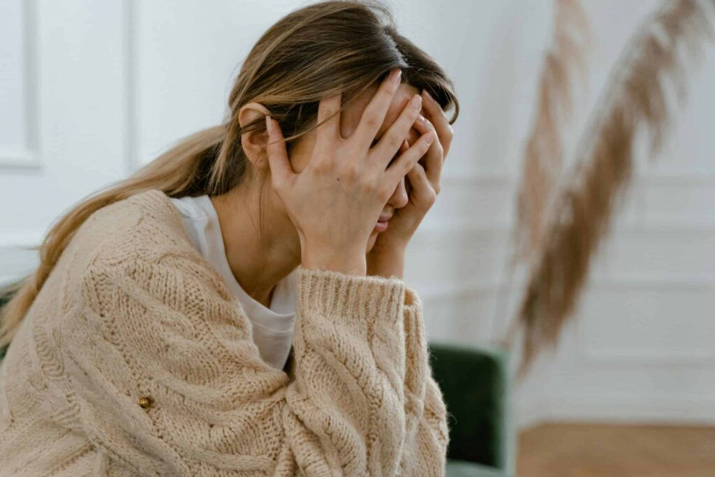 A person in a cream sweater sits with their face in their hands, conveying stress or frustration. The room is softly lit with muted decor, creating a calm backdrop.