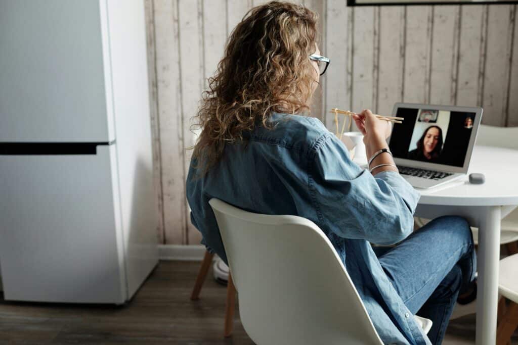 A woman with curly hair, wearing glasses, sits at a white table using chopsticks while video chatting on a laptop.