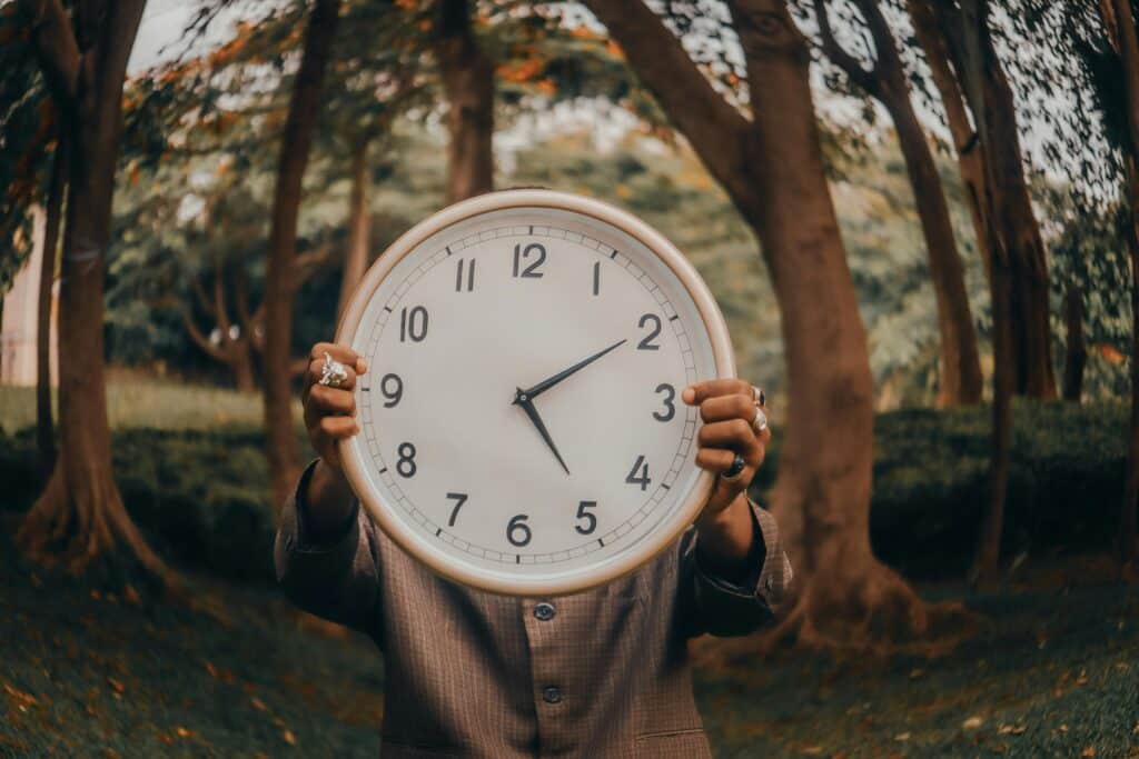 A person in a suit holds a large clock in front of their face, surrounded by trees in a blurred natural setting.