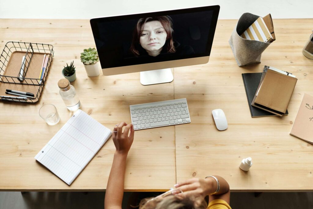 A person's hand holds a pen near a computer setup with a notebook, pens, plants, and a bottle of water on a wooden desk.