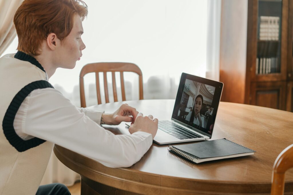A person in a sweater sits at a wooden table, intently viewing a video call on a laptop, with a notepad nearby. Bright light streams in.