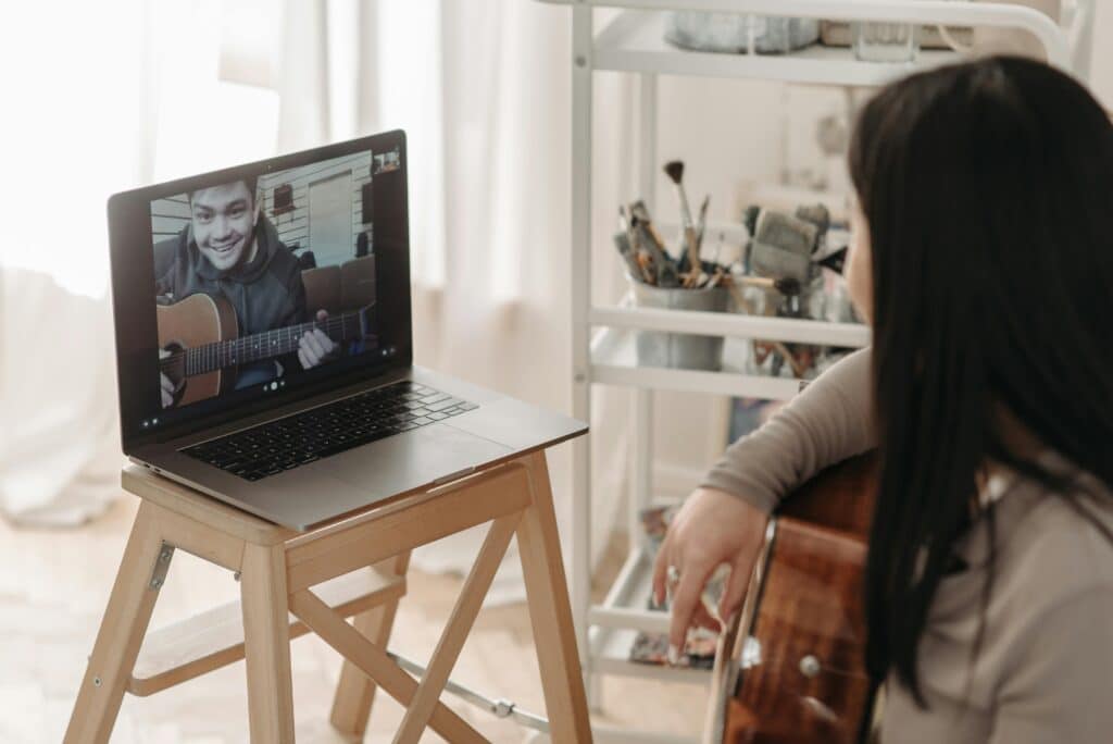 A woman sits with a guitar, focused on a video call with a musician playing another guitar on a laptop. Creative workspace in background.