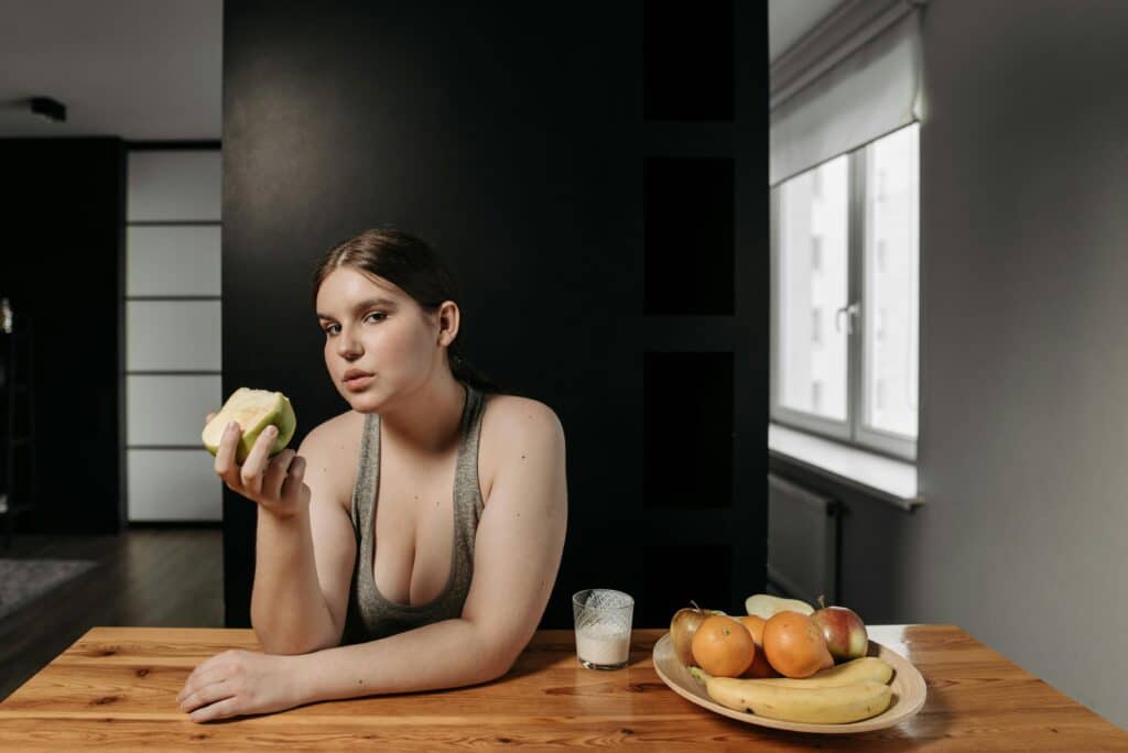 A woman in a gray tank top sits at a wooden table, holding a green apple, with a platter of fruits and a glass of milk nearby.