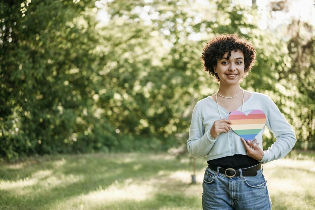 A person stands outdoors, holding a striped rainbow heart, wearing a light sweater, black top, and blue jeans surrounded by greenery.