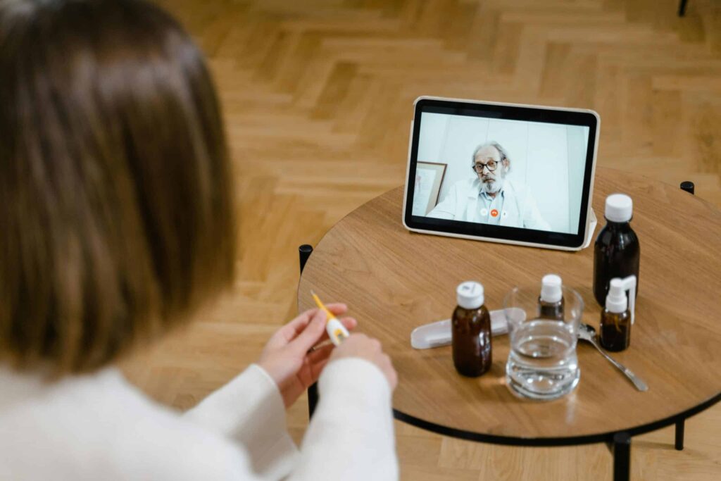 A person sits at a wooden table with medicine bottles, a glass of water, and a tablet displaying a doctor during a teleconsultation.