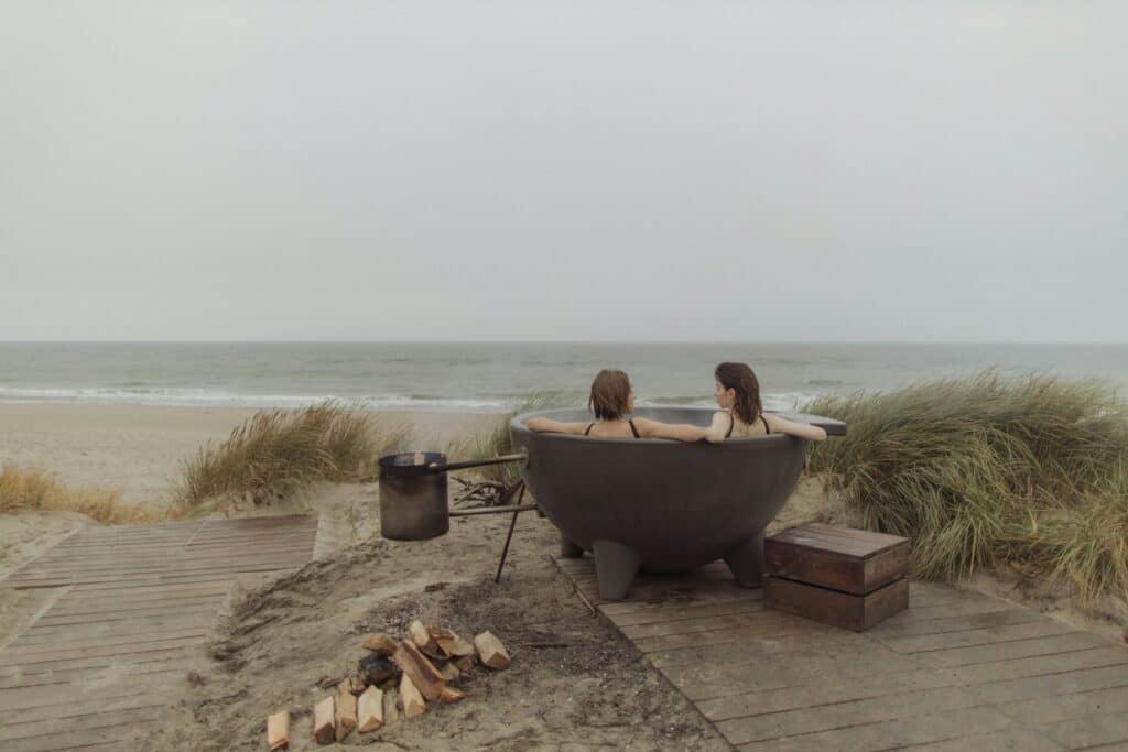 Two women relax in a round hot tub by the beach, with the ocean waves and soft sand in the background. A fire pit sits nearby.