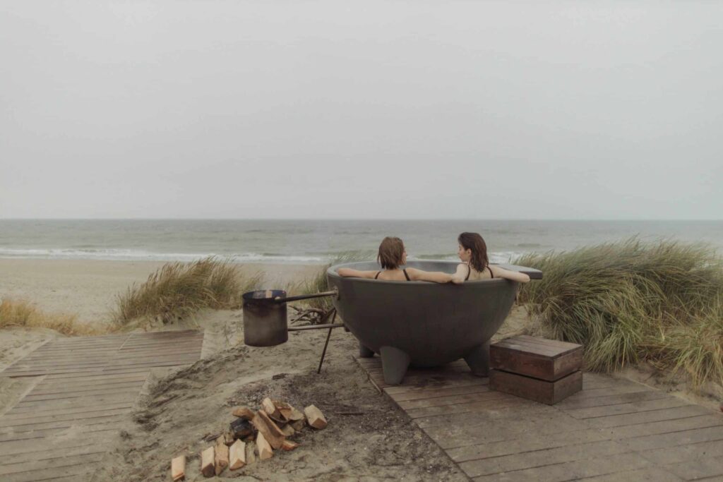 Two women relax in a round hot tub by the beach, with the ocean waves and soft sand in the background. A fire pit sits nearby.