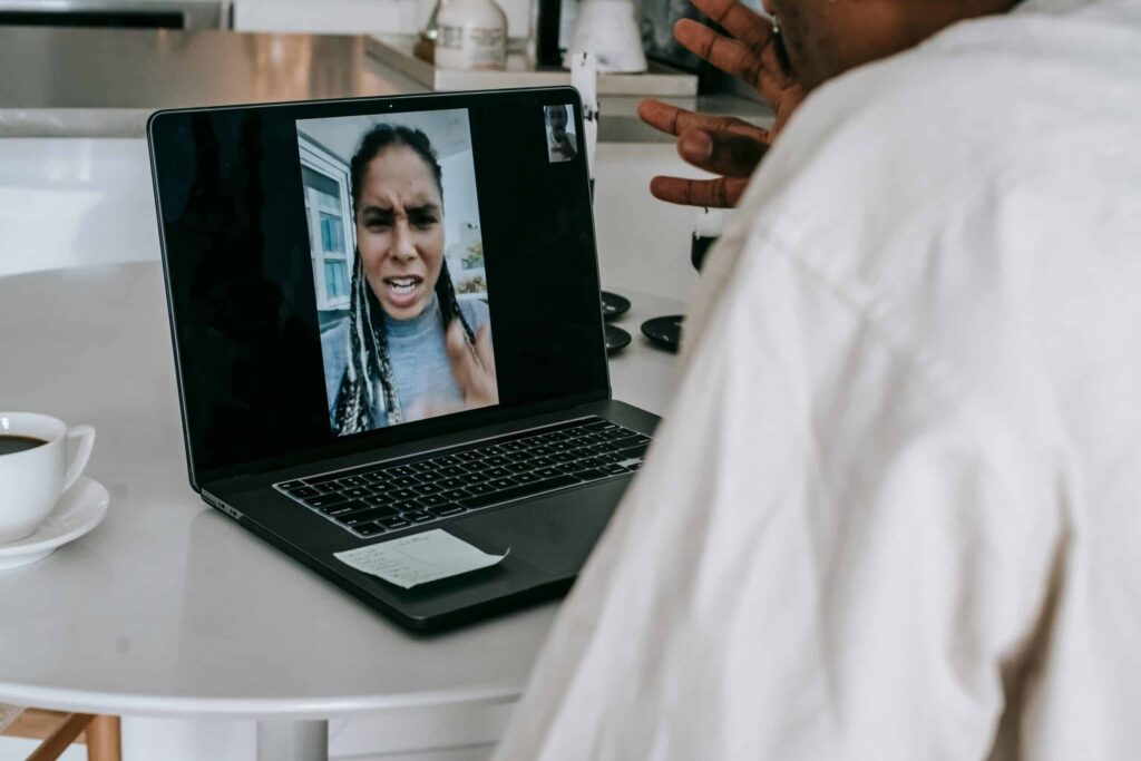A person on a video call, gesturing with one hand, sits at a table with a coffee cup and a laptop displaying the call.