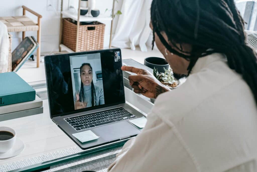 A person with long hair and tattoos intently gestures during a video call on a laptop, with a cup of coffee nearby.