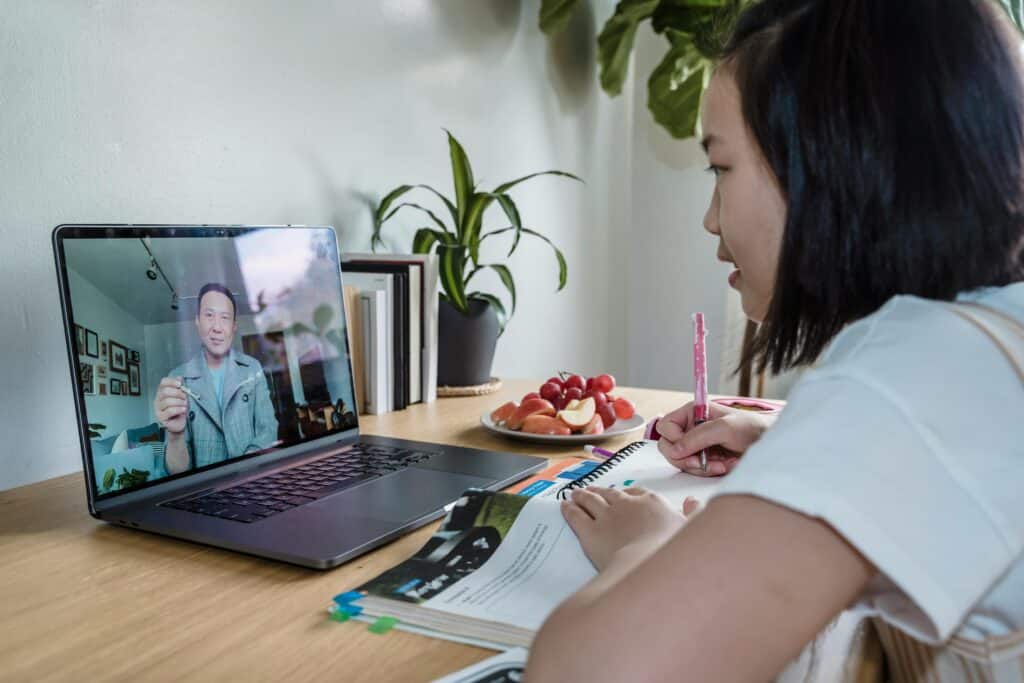 A student writes in a notebook during an online lesson, with a laptop displaying a teacher and a plate of fruits nearby.