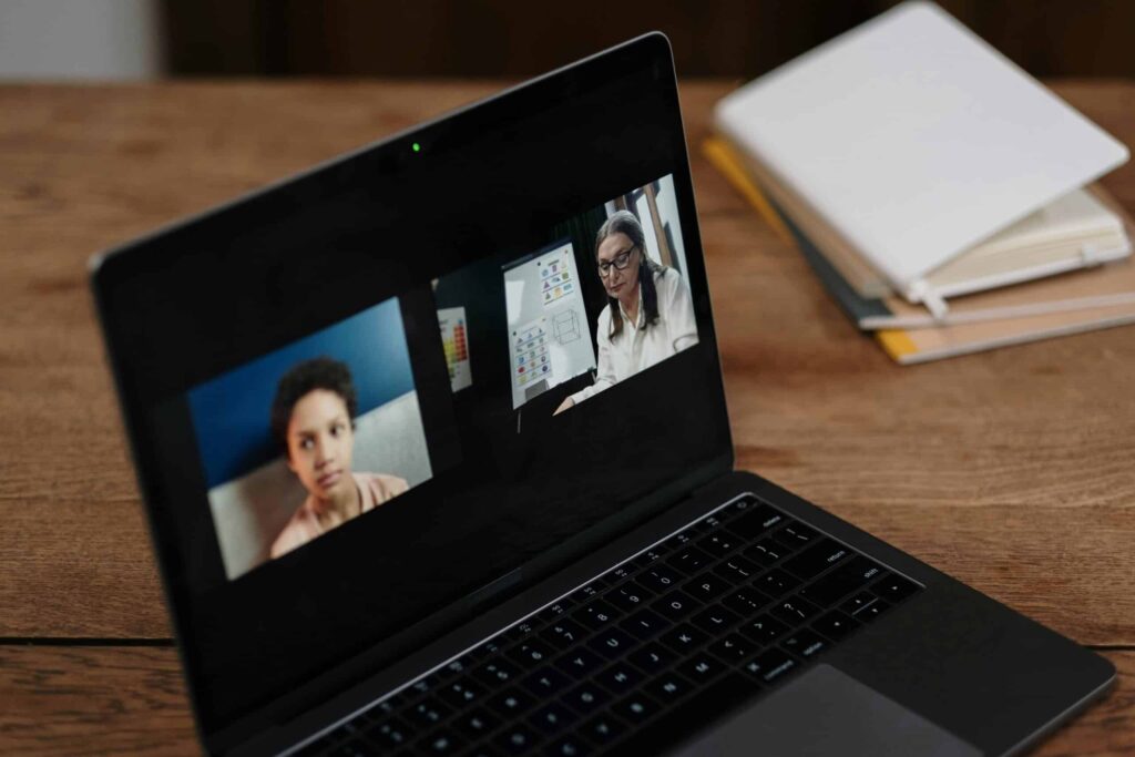 A laptop showing a video call with two participants, one presenting on a whiteboard, and notebooks are visible in the background.