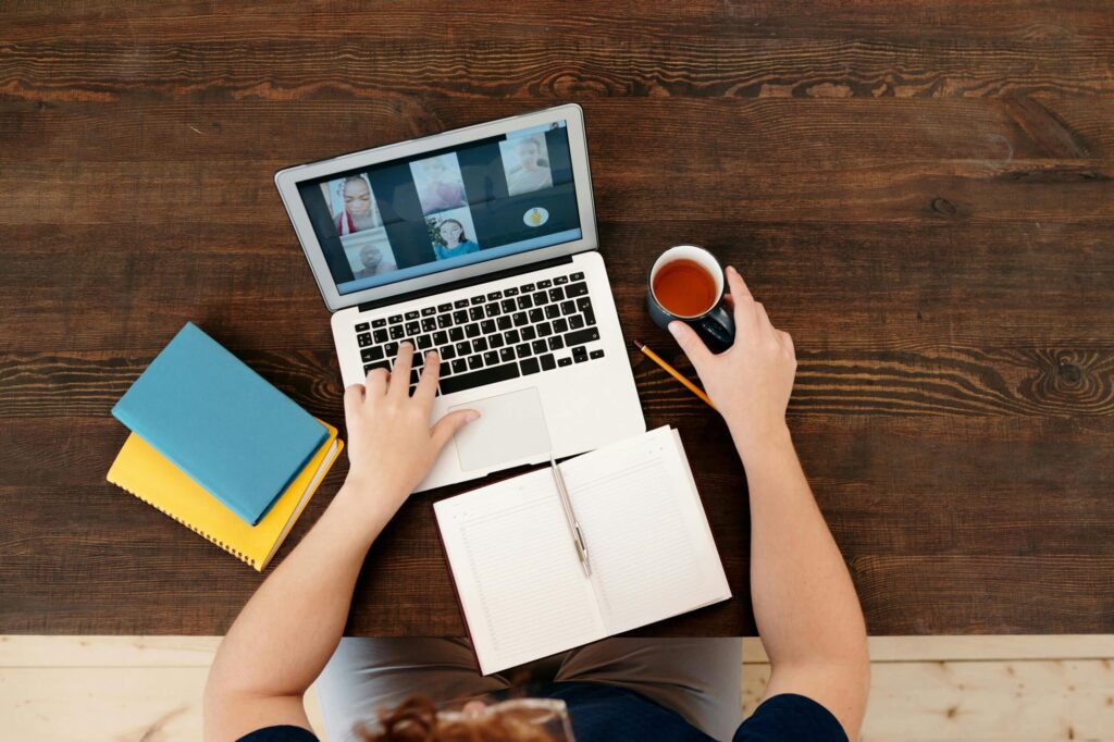 A person sits at a wooden table with a laptop displaying video calls, a notebook, and a cup of tea, actively engaged in work.