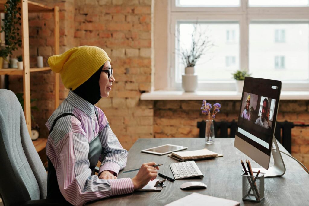 A person in a yellow beanie and striped shirt takes notes while participating in a video call on a computer in a rustic office space.