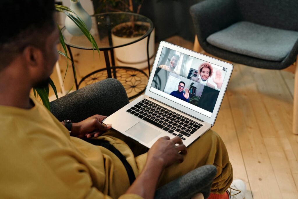 A person sits in a chair, engaged in a video call on a laptop, interacting with multiple participants displayed on-screen.