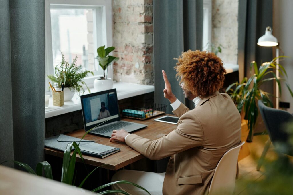 A professional man in a beige suit waves during a video call at a wooden desk surrounded by plants in a bright, modern workspace.